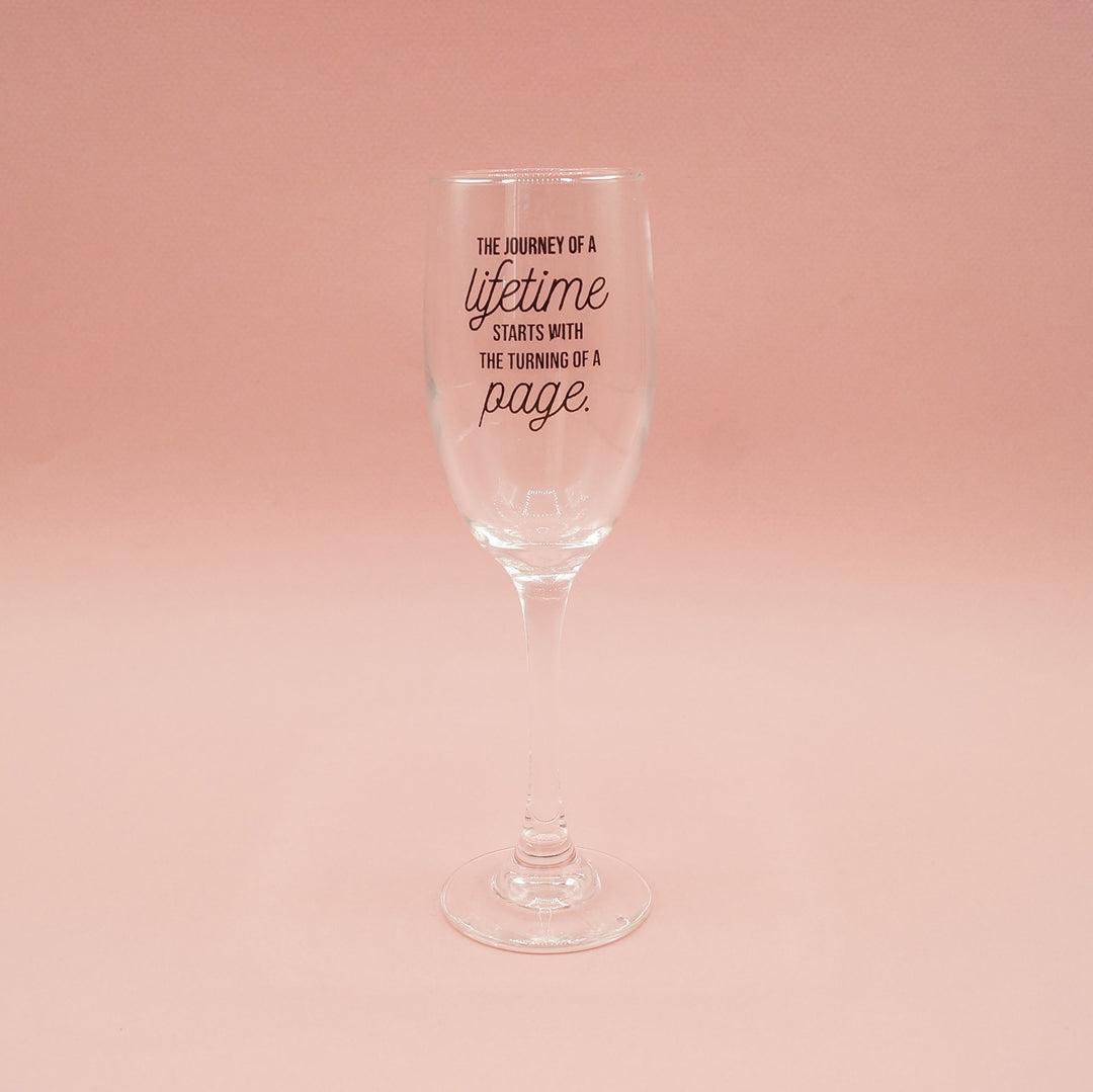 A single champagne flute with the words "The journey of a lifetime starts with the turning of a page" written on the glass sits on a pink background.