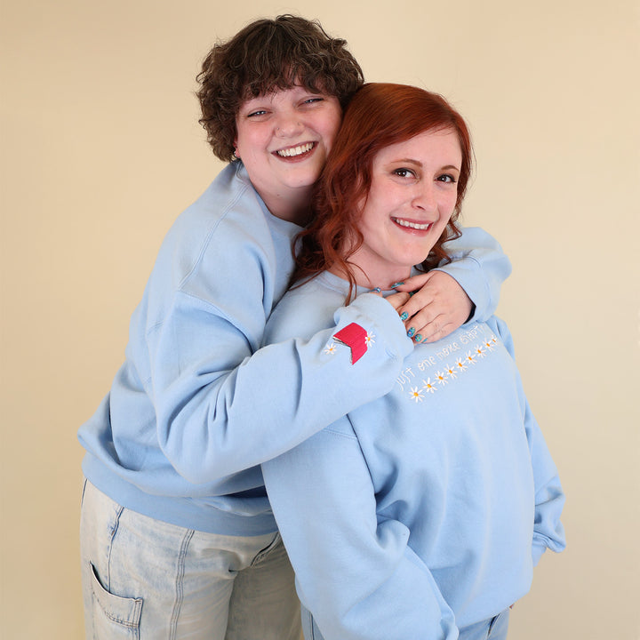 Two women wearing light blue sweatshirts embroidered with 'Just One More Chapter' and daisies on the front and a pink book detail on the sleeve on a beige background