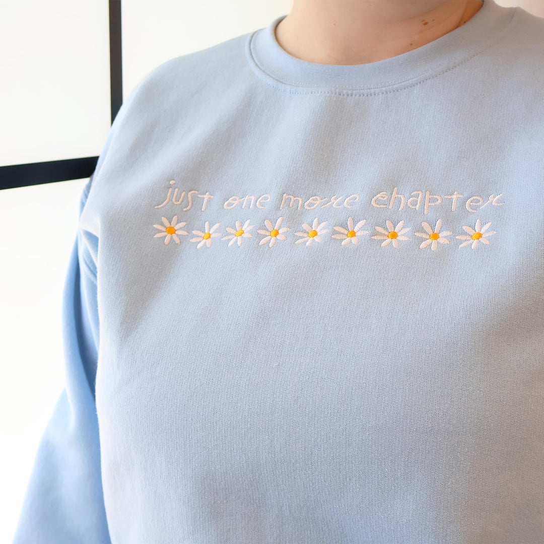 A light blue sweatshirt that's embroidered with 'Just One More Chapter' and daisies on the front in front of a window