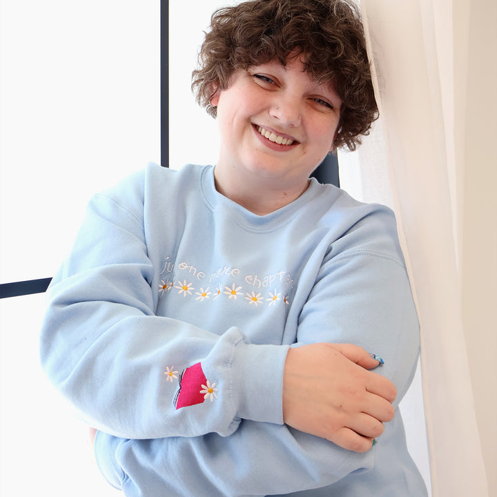 A woman wearing a light blue sweatshirt that's embroidered with 'Just One More Chapter' and daisies on the front and a pink book detail on the sleeve in front of a window