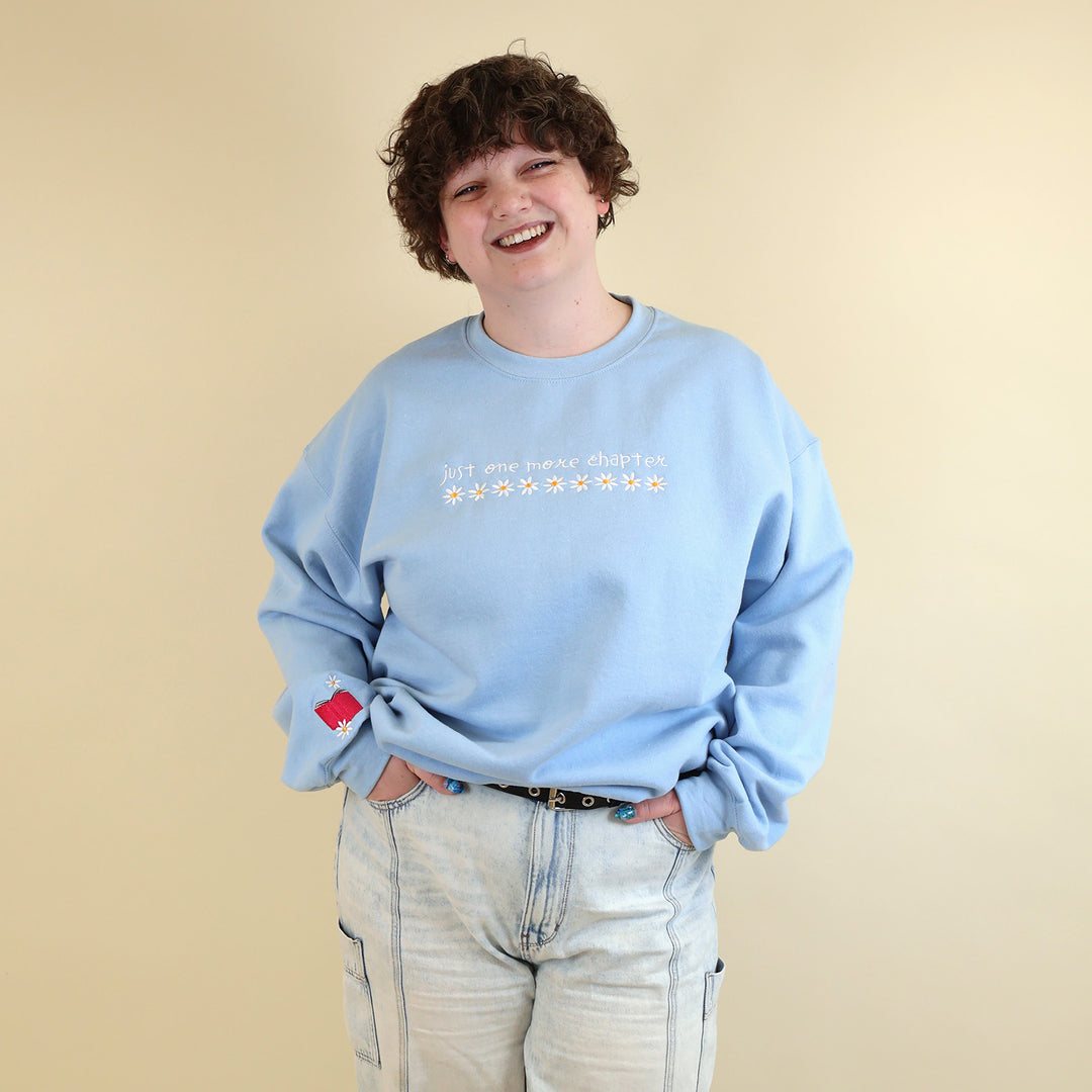 A woman wearing a light blue sweatshirt that's embroidered with 'Just One More Chapter' and daisies on the front and a pink book detail on the sleeve on a beige background