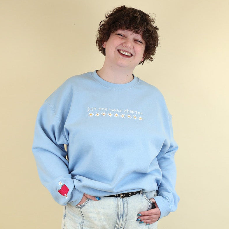 A woman wearing a light blue sweatshirt that's embroidered with 'Just One More Chapter' and daisies on the front and a pink book detail on the sleeve on a beige background
