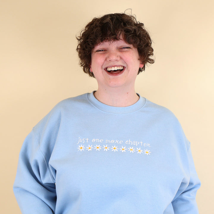 A woman wearing a light blue sweatshirt that's embroidered with 'Just One More Chapter' and daisies on the front and a pink book detail on the sleeve on a beige background