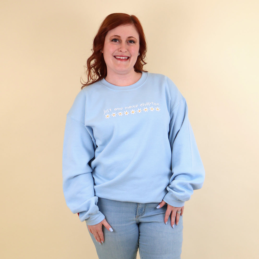 A woman wearing a light blue sweatshirt that's embroidered with 'Just One More Chapter' and daisies on the front and a pink book detail on the sleeve on a beige background