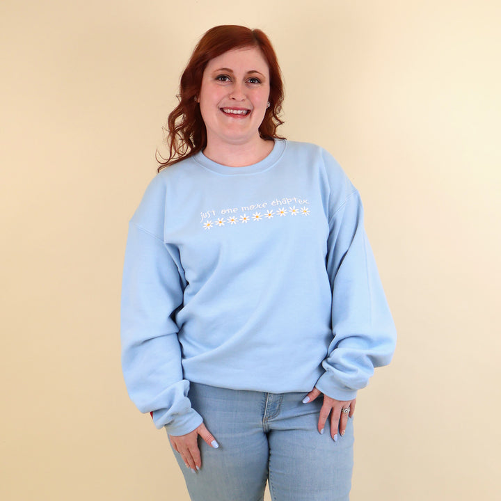 A woman wearing a light blue sweatshirt that's embroidered with 'Just One More Chapter' and daisies on the front and a pink book detail on the sleeve on a beige background