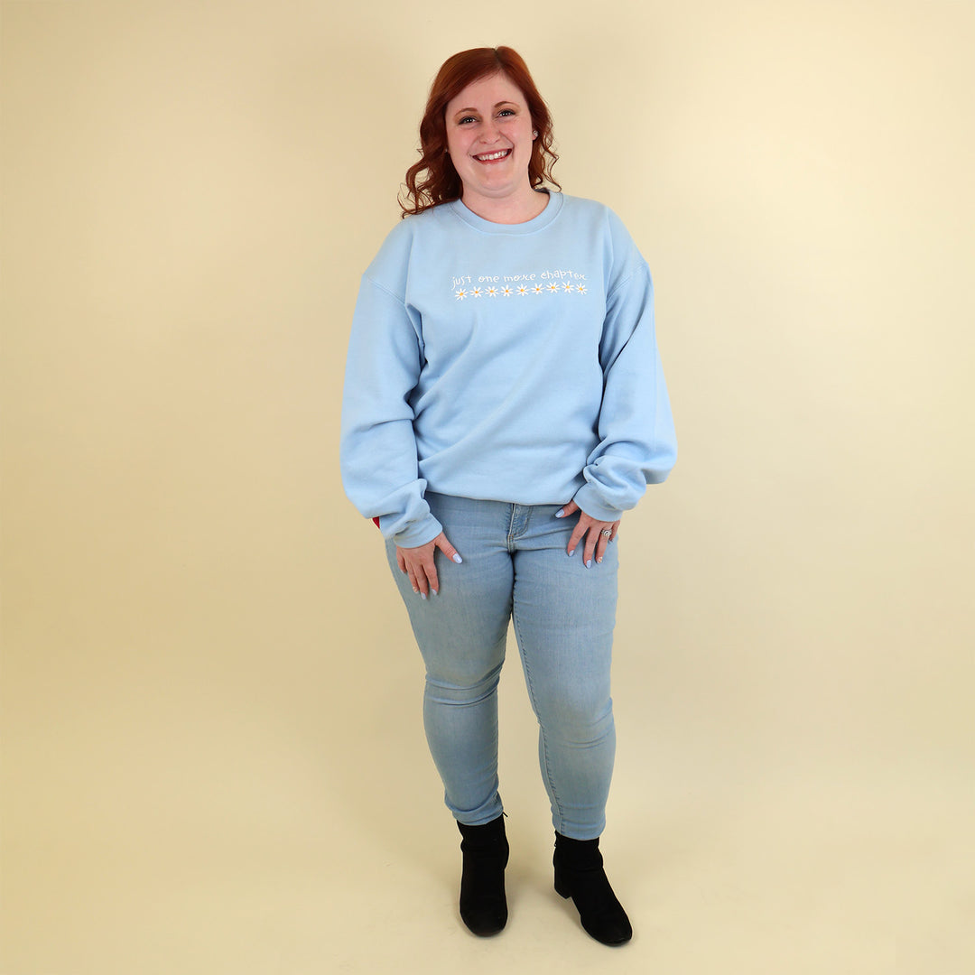 A woman wearing a light blue sweatshirt that's embroidered with 'Just One More Chapter' and daisies on the front and a pink book detail on the sleeve on a beige background