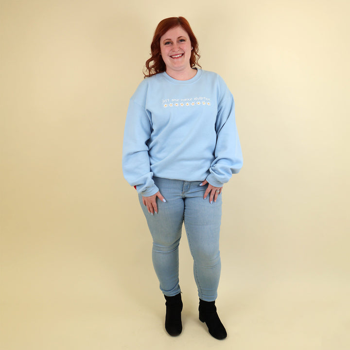 A woman wearing a light blue sweatshirt that's embroidered with 'Just One More Chapter' and daisies on the front and a pink book detail on the sleeve on a beige background
