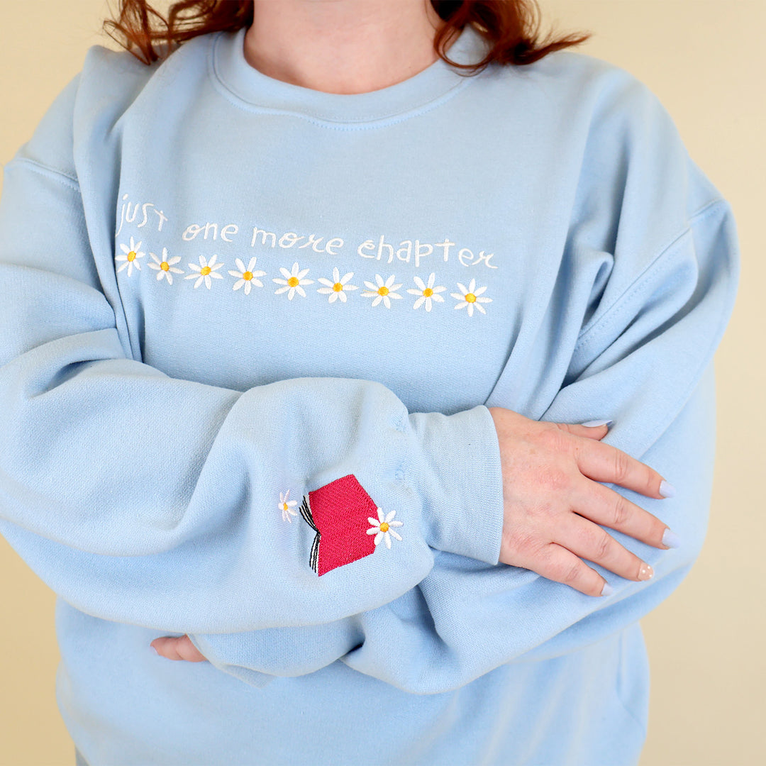 A woman wearing a light blue sweatshirt that's embroidered with 'Just One More Chapter' and daisies on the front and a pink book detail on the sleeve on a beige background