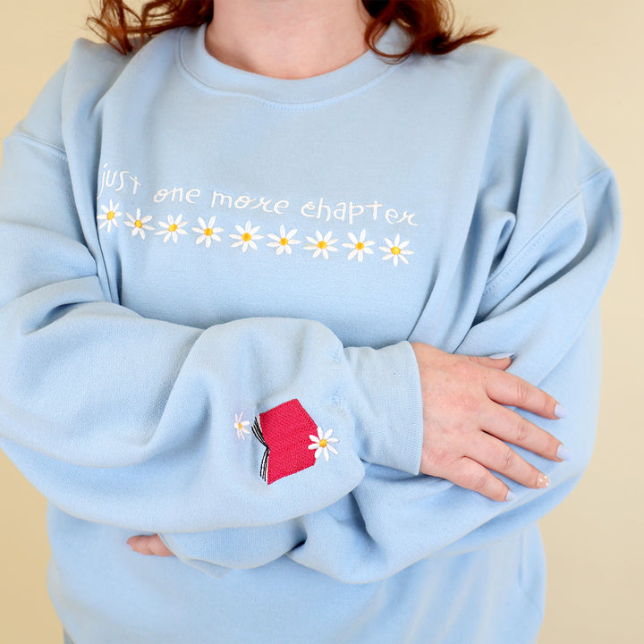 A woman wearing a light blue sweatshirt that's embroidered with 'Just One More Chapter' and daisies on the front and a pink book detail on the sleeve on a beige background