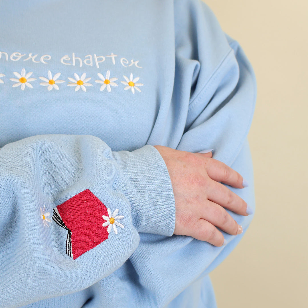 A light blue sweatshirt that's embroidered with 'Just One More Chapter' and daisies on the front and a pink book detail on the sleeve on a beige background