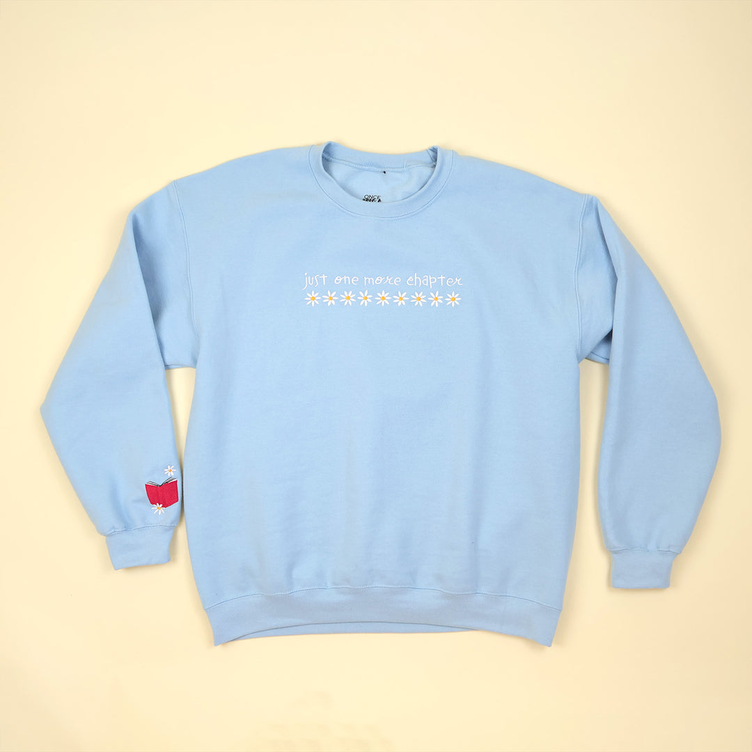 A light blue sweatshirt that's embroidered with 'Just One More Chapter' and daisies on the front and a pink book detail on the sleeve on a beige background