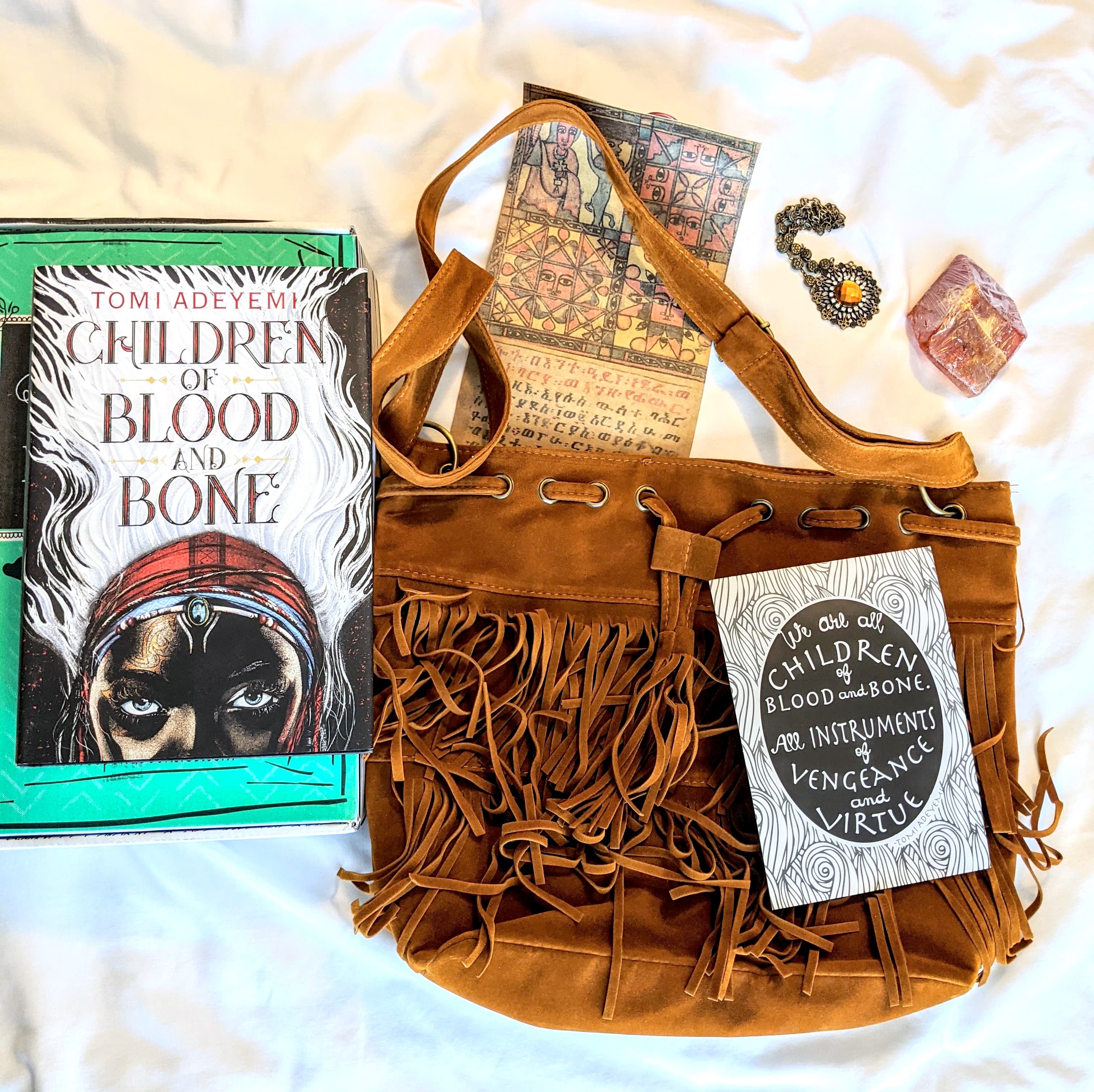 The book Children of Blood and Bone displayed with a fringe bag, a necklace, a gemstone, a map, and a themed quote card.