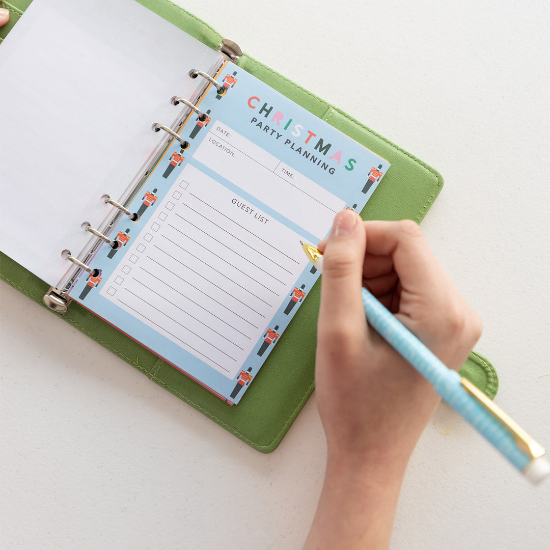 A hand holds a pen over an open green planner. The page inside is labeled Christmas Party Planning with spaces for the Date, Time, Location, and Guest List