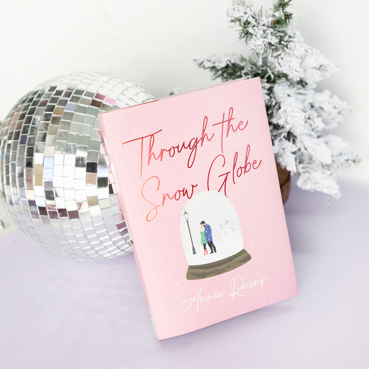 light pink hardcover book "Through the Snow Globe" by Annie Rains standing up against a disco ball and small snowy tree
