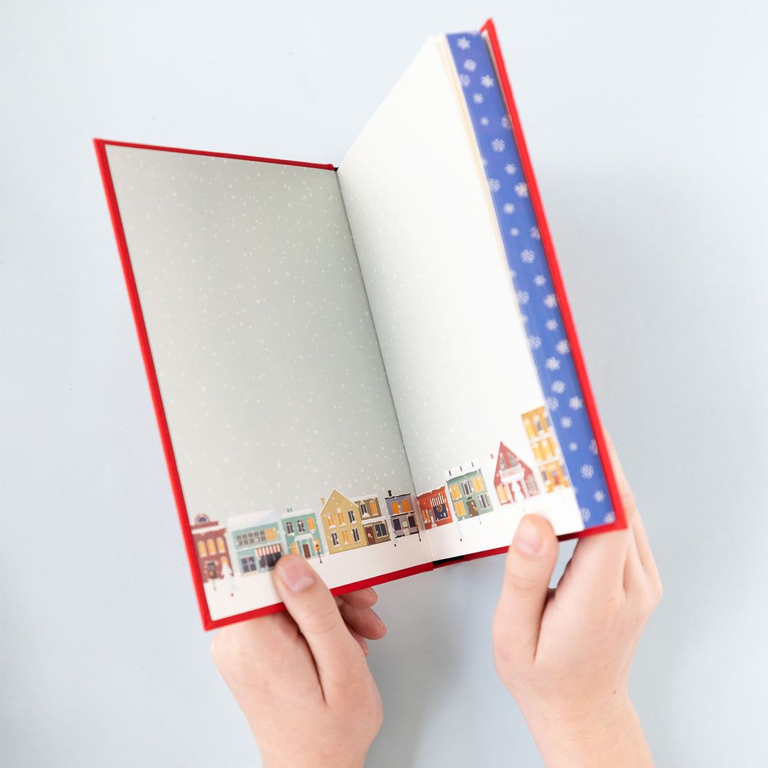 white hands holding a book open, showing end papers of light blue pages with houses along the bottom