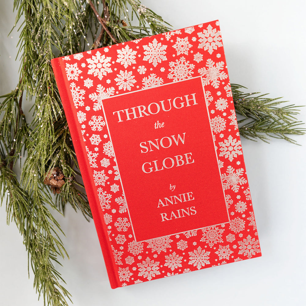 red hardcover book "Through the Snow Globe" by Annie Rains has silver snowflakes around the edges on the cover, sitting on a tree branch
