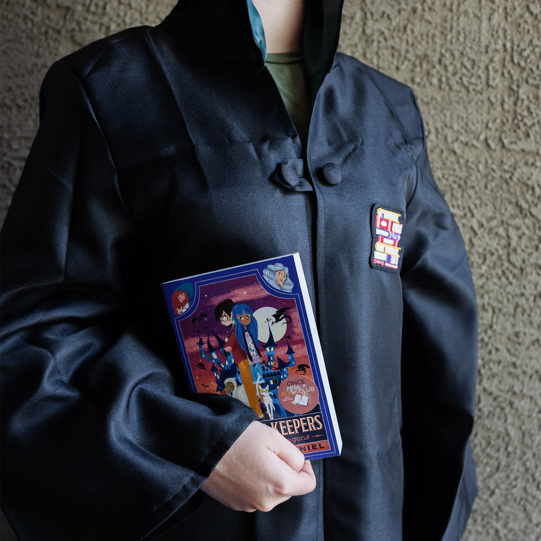 A child wears a black cloak with a bookstack patch on the front right, while holding a paperback book in hand.