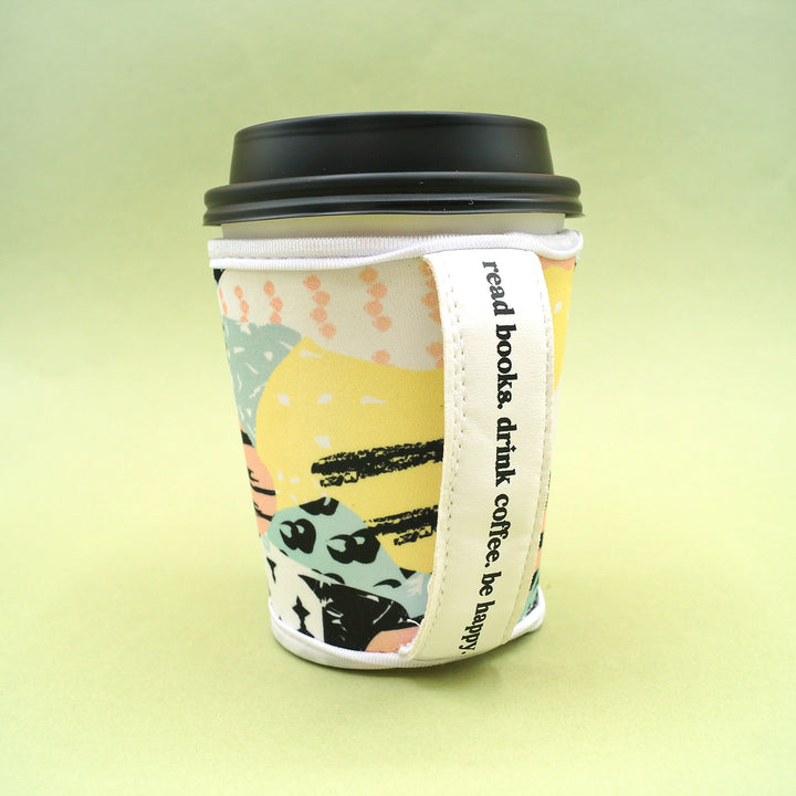 a coffee cup cozy sits on a green background with a coffee cup in it. The quote "read books, drink coffee, be happy" is printed on the handle
