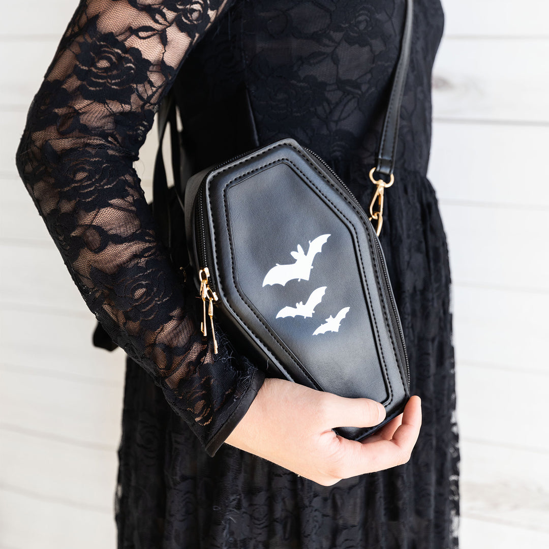 A white woman wearing a black lace dress cradles a black coffin-shaped purse with white bats on it to her side