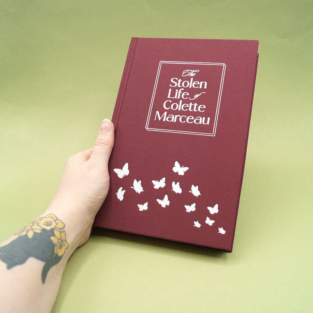 Maroon book titled 'The Stolen Life of Colette Marceau' held by a hand on a green background