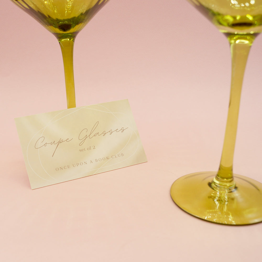 A close up the tag for the set of 2 coupe glasses by Once Upon a Book Club. The stems of the two green coupe glasses are visible. On a pink background.