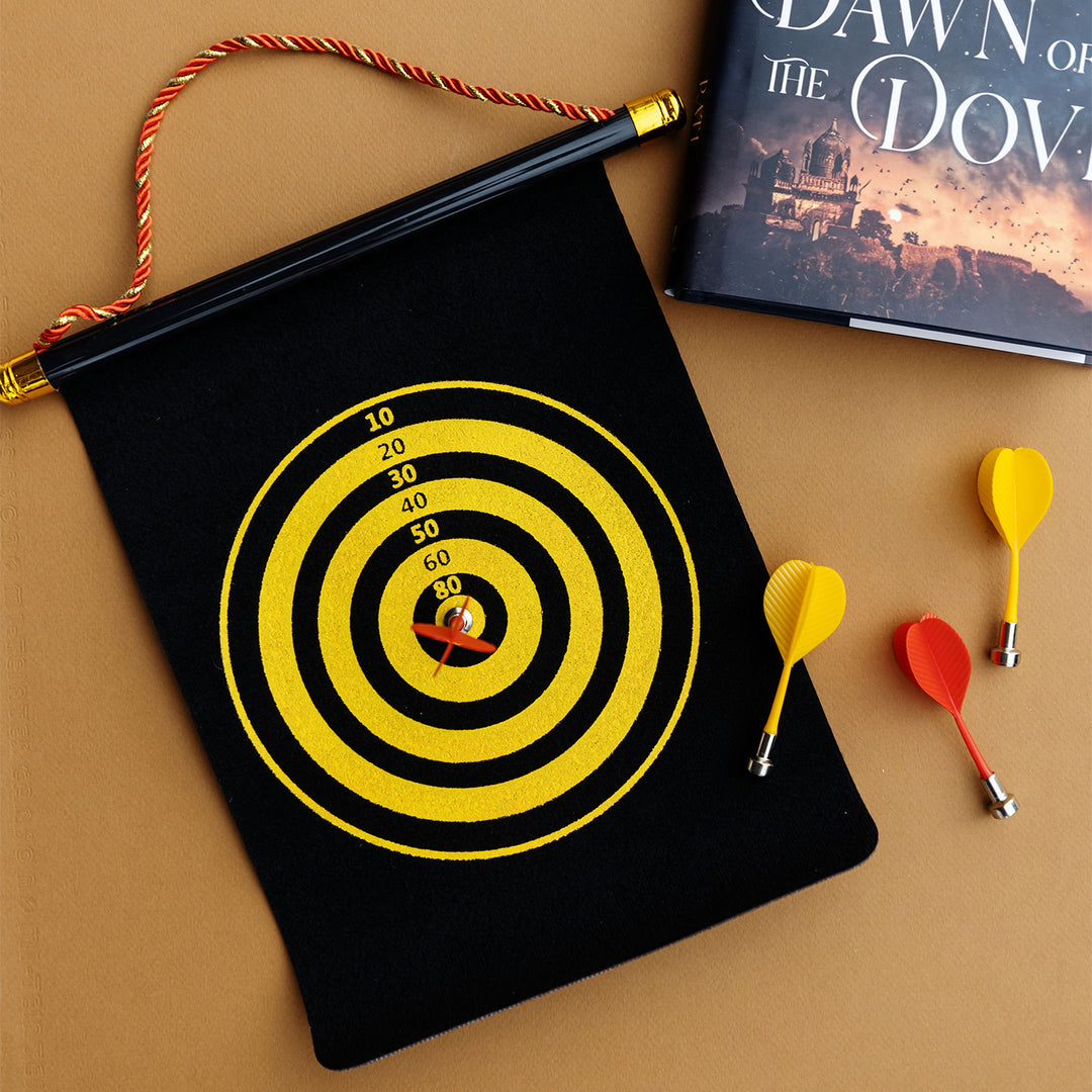 a roll-up black dartboard with two yellow darts and two red darts. The scoring circle is in alternating colors of yellow and black. A hardcover edition of Night of the Raven, Dawn of the Dove is next to the dart board.