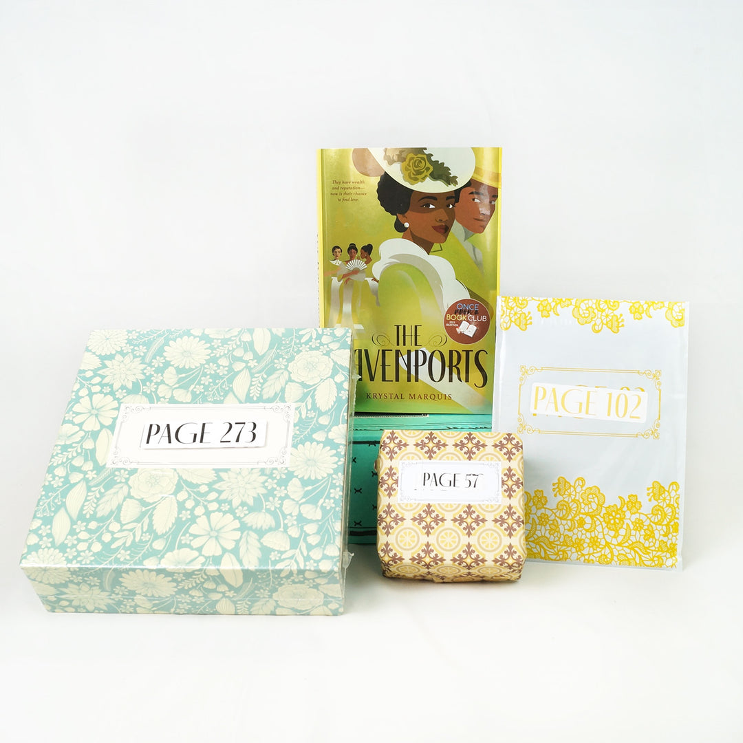 A hardcover edition of The Davenports sits on a green Once Upon a Book Club box. In front of the green box are a light green floral box, yellow square box, and yellow and white folder. The boxes and folder all have page numbers.