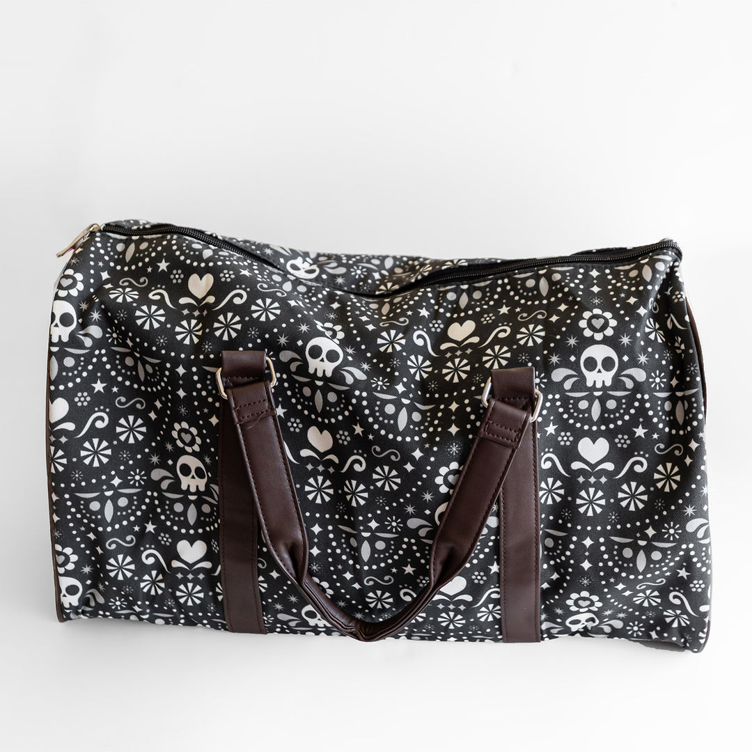 A black duffel bag with dark brown straps. The duffel bag has a pattern of white skulls, hearts, and stars on it.
