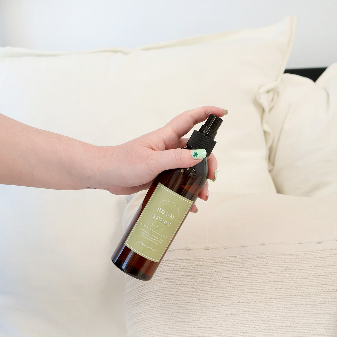 A hand holds out a bottle of Lemon Verbena Room Spray over white sheets.