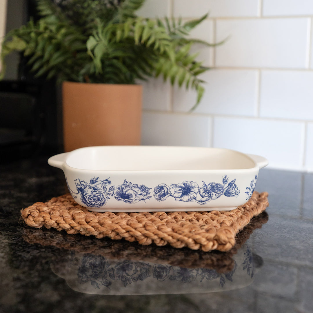 A white baking dish with a blue floral pattern on the side sits on a woven pot holder on a black counter.