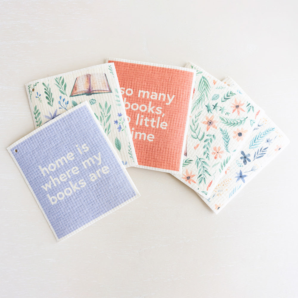 A collection of Swedish dishcloths in pastel tones featuring flowers and books. The top towel reads "home is where my books are"