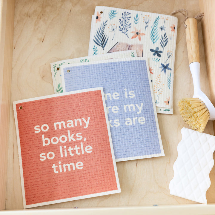 A collection of Swedish dishcloths in pastel tones featuring flowers and books in a wooden drawer. The top towel reads "So many books, so little time."