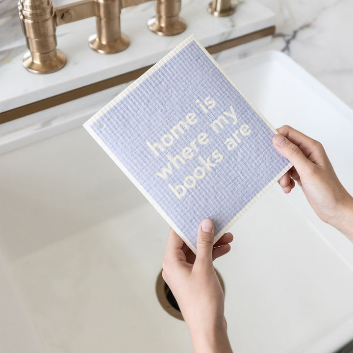A set of hands holds a Swedish dishtowel over a sink. The towel says "home is where my books are"