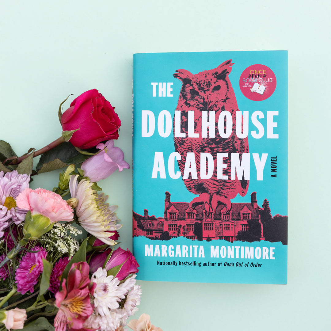 A hardcover edition of The Dollhouse Academy by Margarita Montimore sits next to a bouquet of flowers.