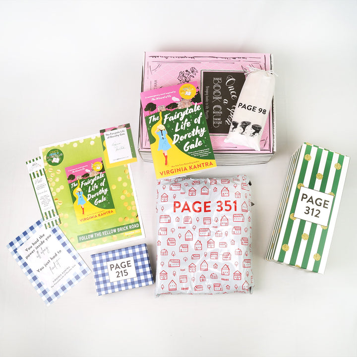 a paperback copy of "The Fairytale Life of Dorothy Gale" by Virginia Kantra next to a white drawstring bag with black tornados sits on a pink box. In front are paper items (quote card, bookmark, bookclub kit, signature card), a blue and white checkered square box, a white bag with red houses, and a green/white striped rectangular box. The boxes and bags all have page numbers.