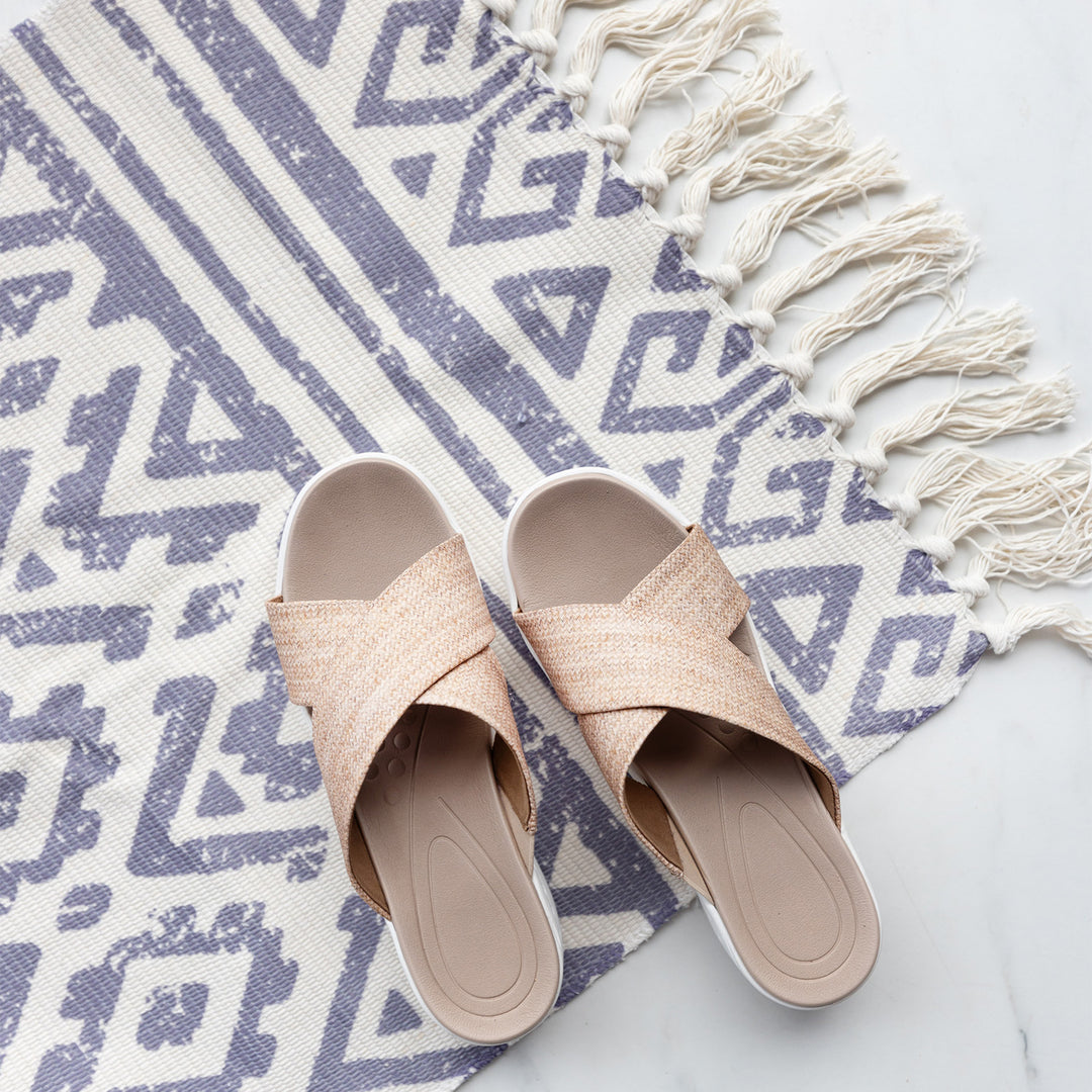 A cream and grey toned floor mat printed with geometric patterns. Cream colored tassels hang off the end and a pair of tan sandals sit on top of the mat.