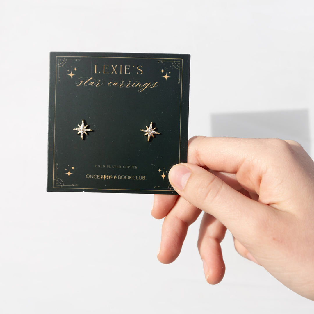 A hand holds a square card labeled Lexie's Star Earrings with two star-shaped earrings attached. At the bottom it says Gold Plated Copper Once Upon a Book Club.