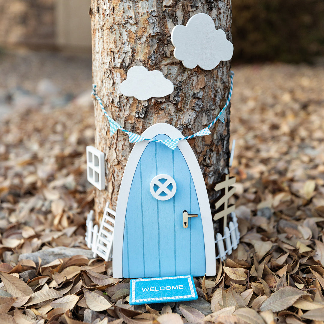 A light blue and white fairy door is attached to the trunk of a tree. White clouds are above it and a white picket fence is on each side. A small light blue welcome mat is in front.