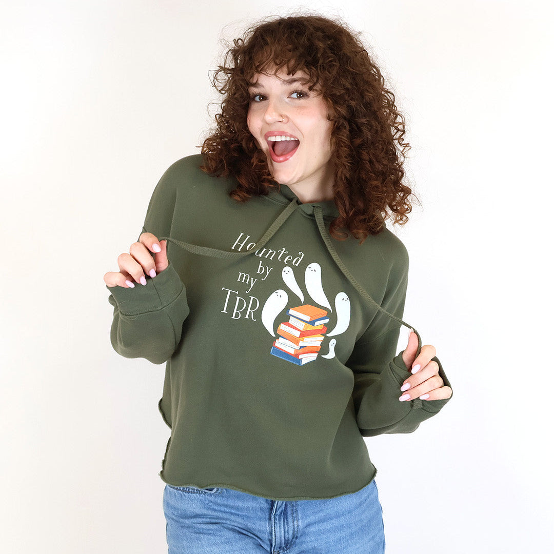 A woman with curly brown hair wearing an olive green hoodie with a 'Haunted by my TBR' graphic and blue jeans, posing against a white studio background.
