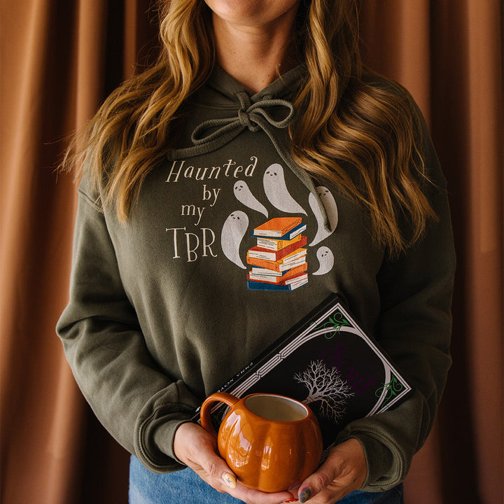 A woman wearing an olive green hoodie with a graphic that reads 'Haunted by my TBR' and shows ghost illustrations next to a stack of books, holding an orange mug in front of a brown curtain background.