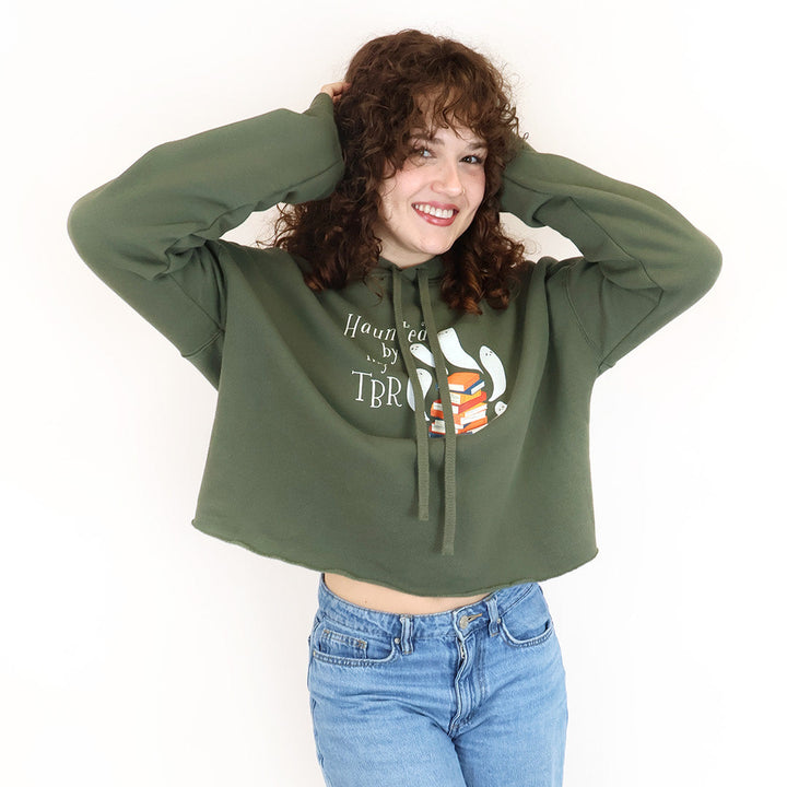 A woman with curly brown hair wearing an olive green cropped hoodie with a 'Haunted by my TBR' design and blue jeans, posing against a plain white background.