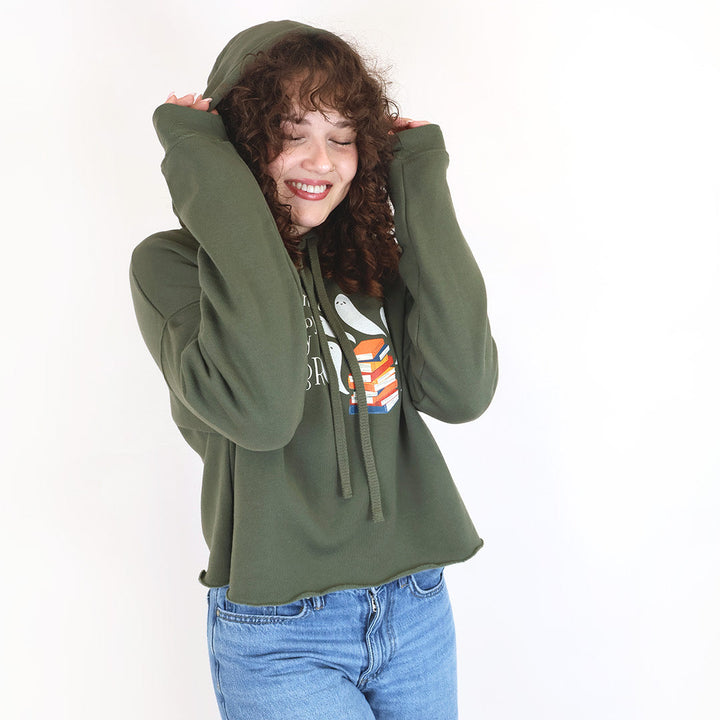 A woman with curly brown hair wearing an olive green cropped hoodie with a 'Haunted by my TBR' design and blue jeans, posing against a plain white background.