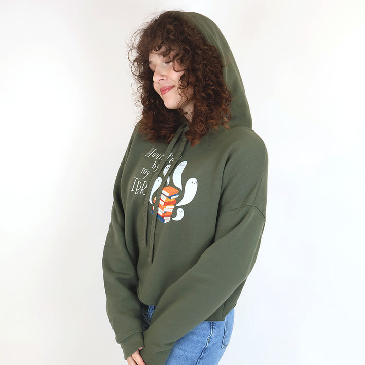 A woman with curly brown hair wearing an oversized olive green hoodie that reads "Haunted by my TBR" with a graphic of white cartoon ghosts and a colorful stack of books, paired with blue jeans, set against a plain white background.