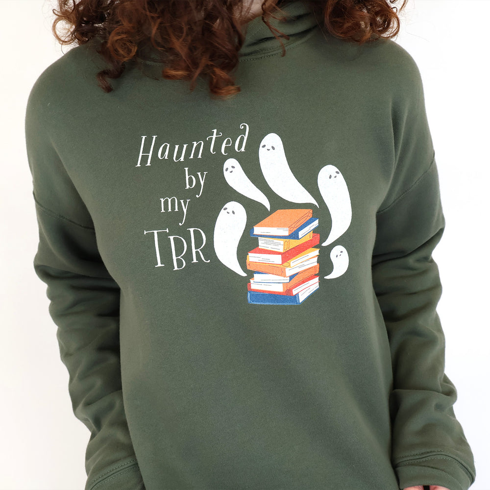 A woman wearing an olive green hoodie with a graphic on the chest that reads 'Haunted by my TBR' in white script, accompanied by white cartoon ghosts and a stack of colorful books.