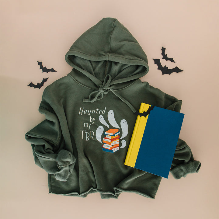 An olive green hoodie with a 'Haunted by my TBR' graphic, styled in a flat lay with a blue and yellow notebook, black bat cutouts, and a beige background.