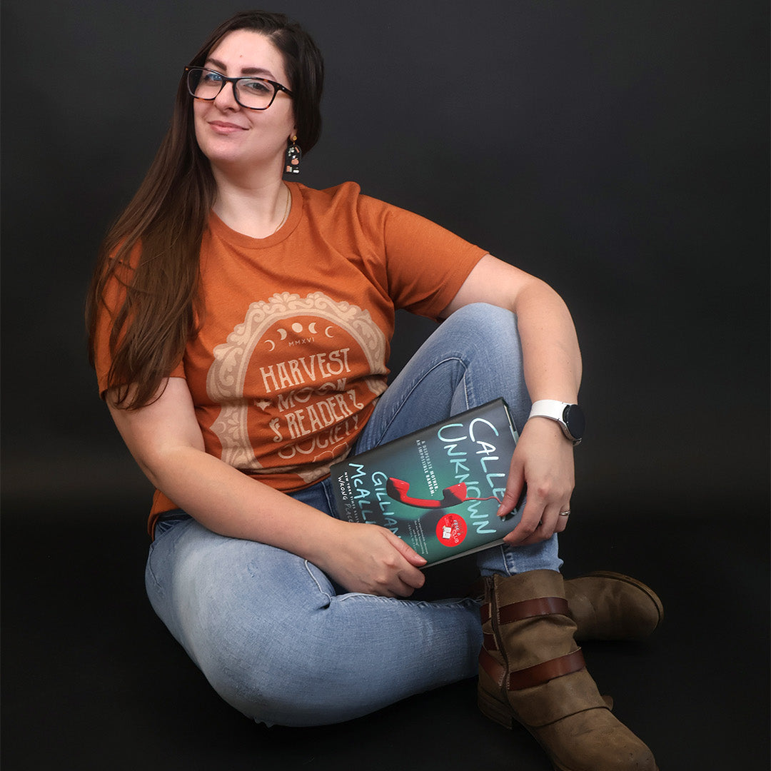 A woman with long brown hair and glasses sits cross-legged against a dark studio background, wearing an orange graphic T-shirt that reads "Harvest Moon Reader Society", light blue jeans, and brown boots while holding a book.