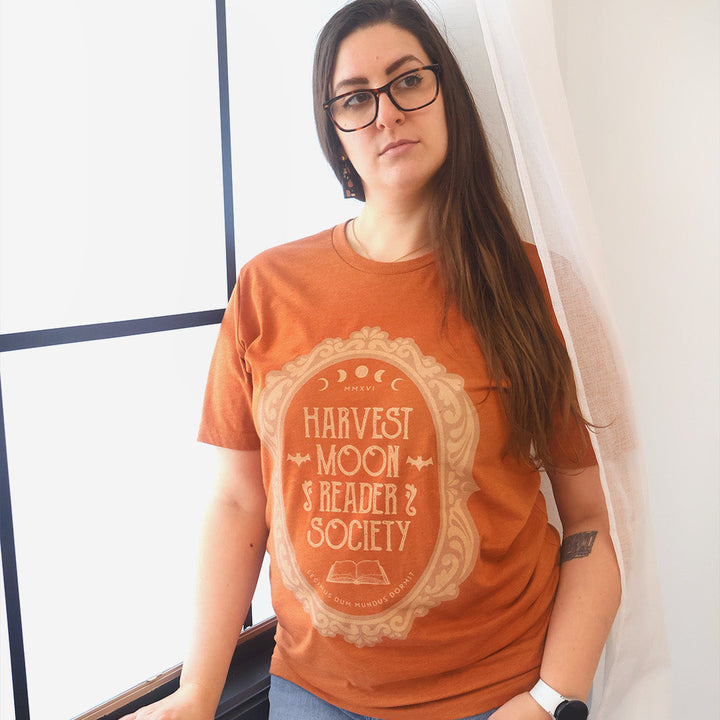 A woman with long brown hair and glasses wearing a rust-orange graphic t-shirt with a cream-colored celestial print that reads 'Harvest Moon Reader Society'. She is wearing blue jeans and standing next to a white curtain.