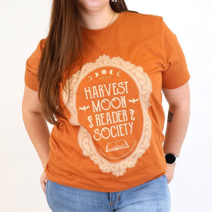 A woman wearing a burnt orange graphic t-shirt with a cream-colored ornate frame and the text 'Harvest Moon Reader Society' on the front, paired with light blue jeans against a plain white background.