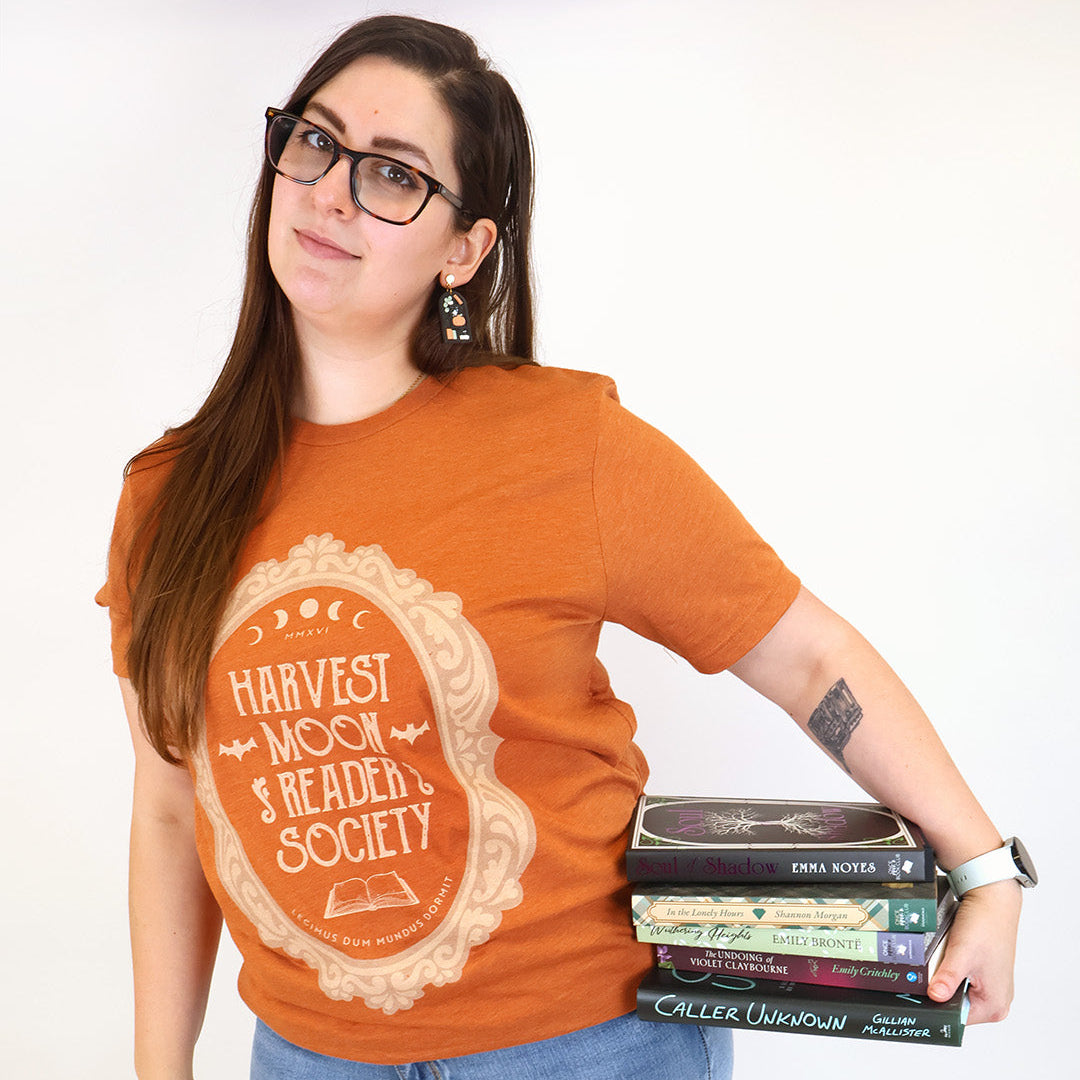 A woman wearing a burnt orange graphic t-shirt with a cream-colored 'Harvest Moon Reader Society' emblem, paired with blue jeans and holding a stack of books against a plain white background.
