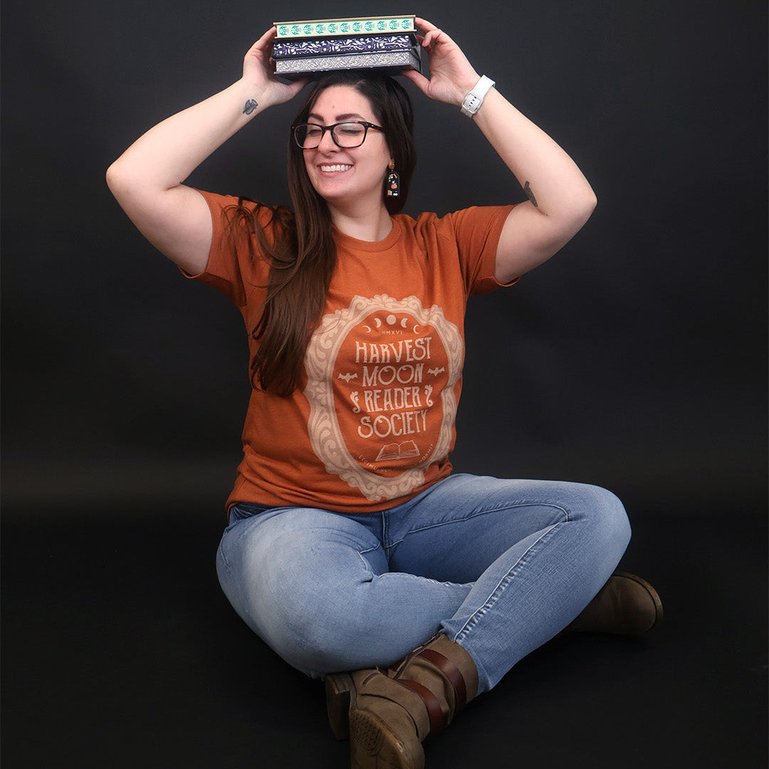 A woman wearing a rust-orange graphic t-shirt with a 'Harvest Moon Reader Society' design, sitting cross-legged against a black studio background while balancing a book on her head.